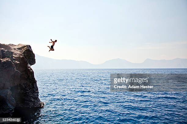 young man jumping off of cliff into crater lake on sunny day, crater lake national park, oregon, usa - roccia foto e immagini stock