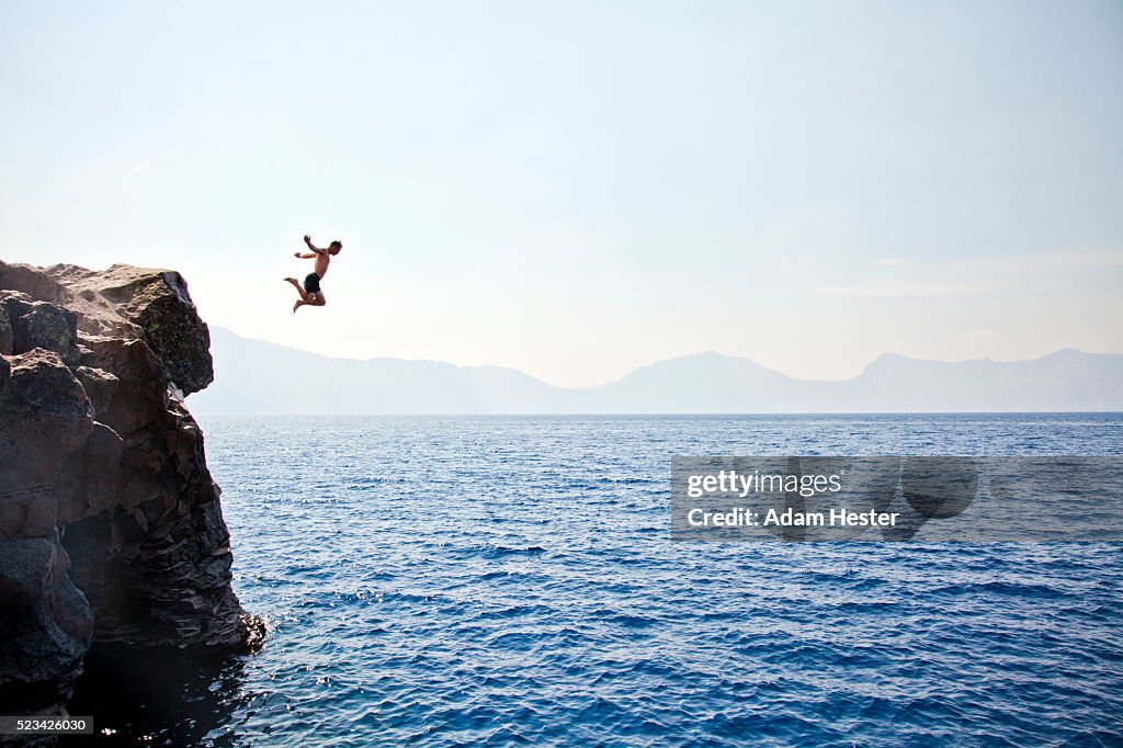 Young man jumping off of cliff into Crater Lake on sunny day, Crater Lake National Park, Oregon, USA