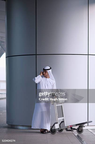 man wearing traditional clothing standing with airport luggage trolley, using mobile phone - arabic people ストックフォトと画像