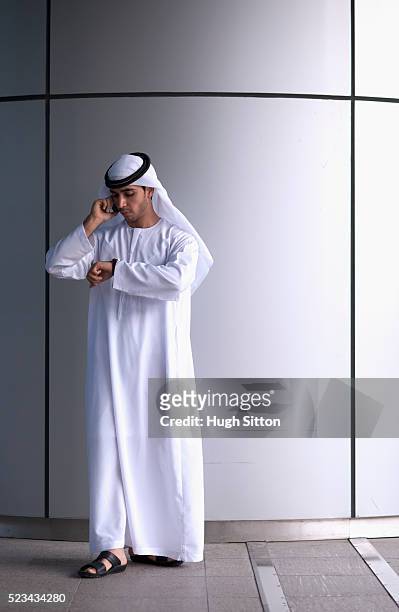 man wearing traditional clothing checking time and using mobile phone - arab businessman stock pictures, royalty-free photos & images