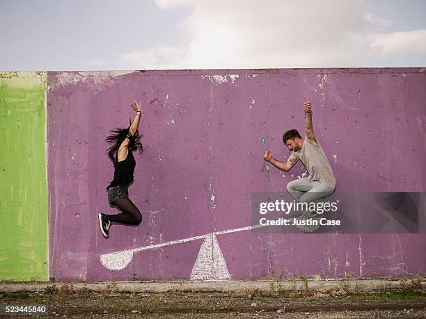 young man jumping on painted seesaw to push young woman up - painted wall stockfoto's en -beelden