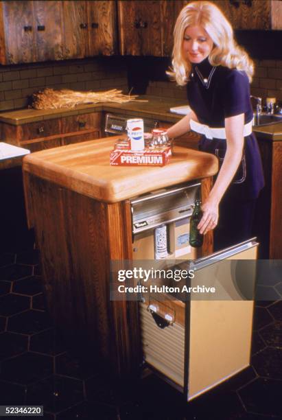 Blonde woman in a blue pantsuit with white stitching and belt places household food packaging in a Whirlpool brand trash compactor in a kitchen,...