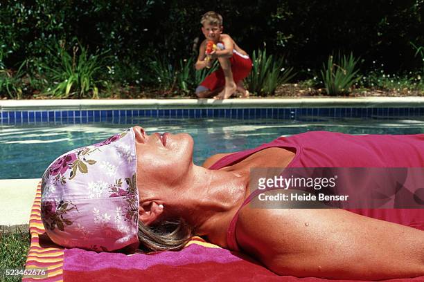 boy at swimming pool aiming with water pistol at sunbathing woman - misbehaving children stock pictures, royalty-free photos & images