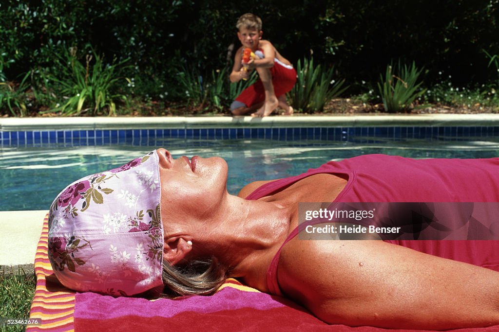 Boy at swimming pool aiming with water pistol at sunbathing woman