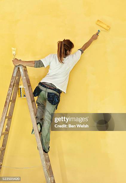 young woman painting wall with yellow paint - woman on ladder painting stock pictures, royalty-free photos & images