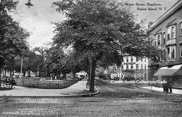 Postcard showing the Staten Island Savings Bank on corner, shops, Washington Park across street, brick road with trolley track, men standing on...