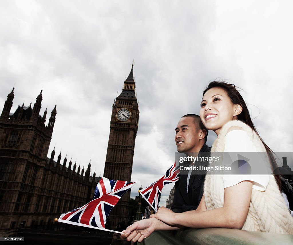 Smiling Asian tourist couple waving small Union Jacks in front of Houses of Parliament, London, England, UK