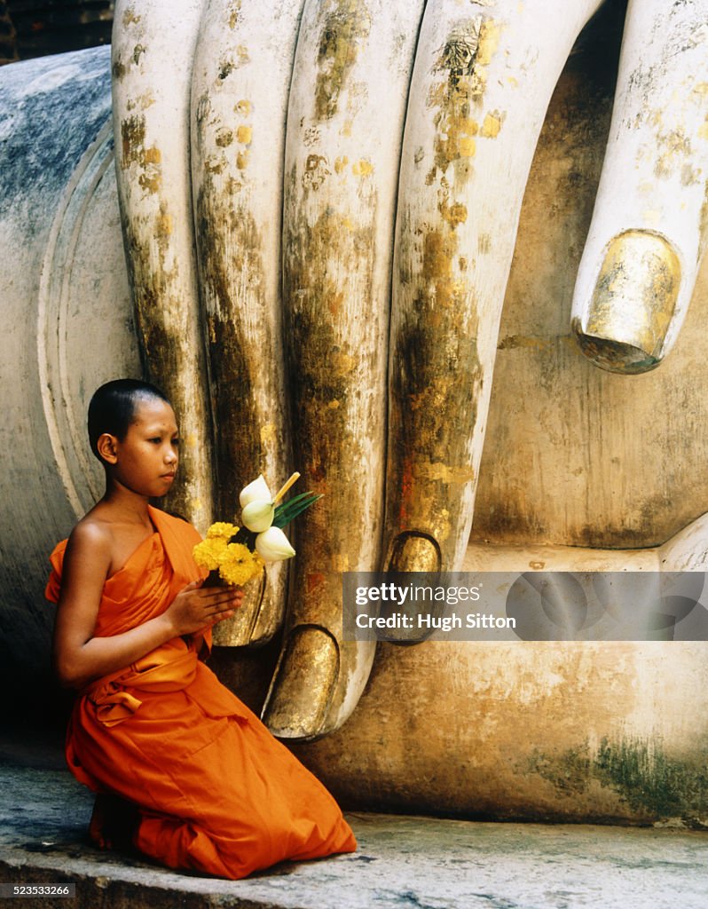 Novice Buddhist Monk Kneeling Next to Fingers of Statue