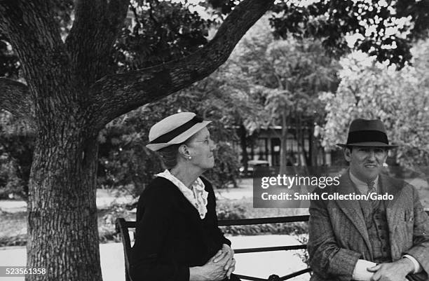 Photograph of a woman and a man sitting on a metal park bench, the woman is wearing a dark dress and a hat, the man is wearing a grey suit and tie...