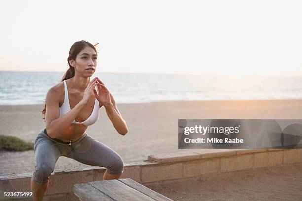 young woman doing squat jumps on beach - hockend stock-fotos und bilder