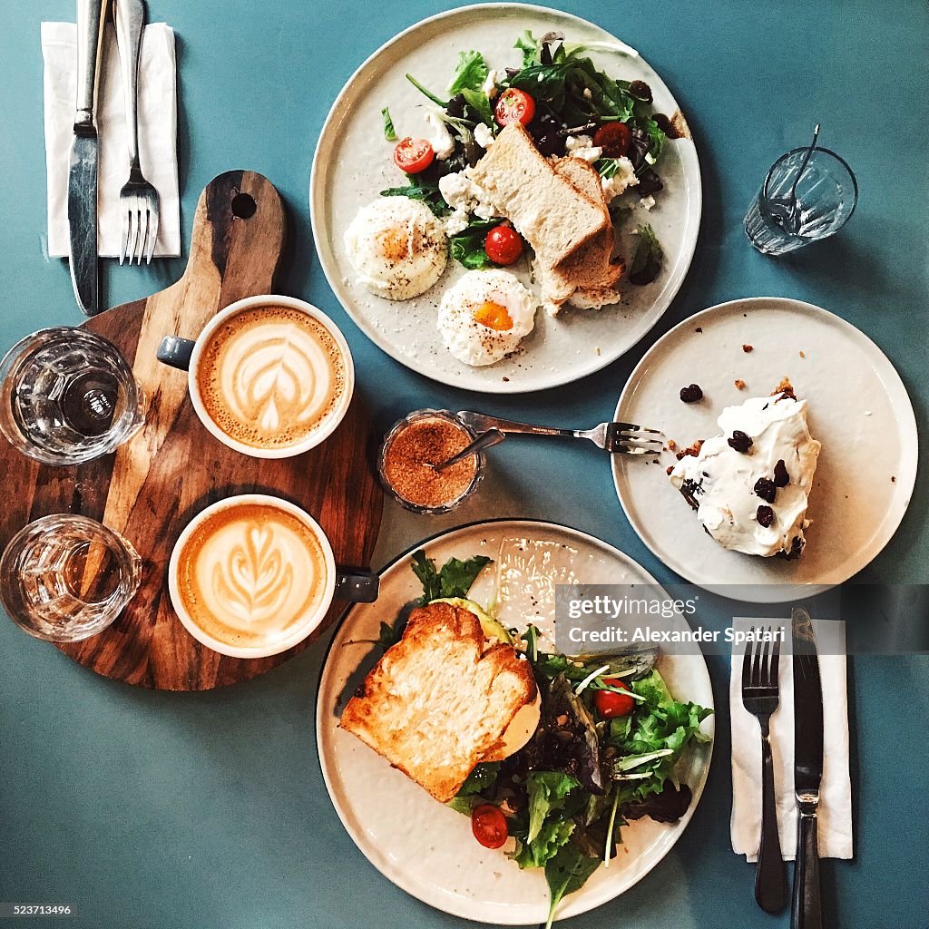 Brunch for two people with avocado toast, fried egg, salad, cappuccino and carrot cake served on the table, high angle view