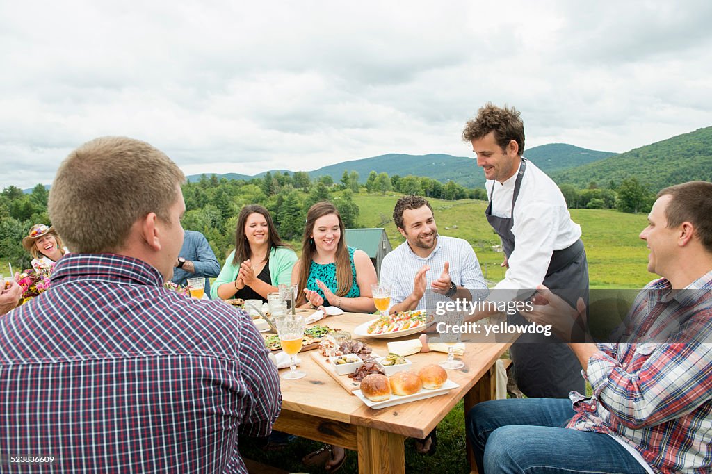 Mid adult man in apron, serving plate of food to family members at table, outdoors