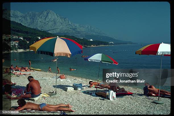 Tourists sunbathe on an umbrella dotted beach below green hills and a rocky mountain at Brela, Yugoslavia. | Location: Brela Shores, Yugoslavia.