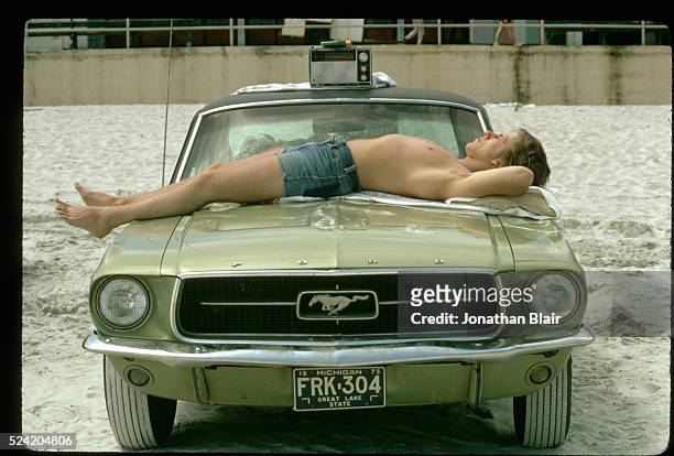 Young man sunbathes on the hood of his green Ford Mustang on Daytona Beach.
