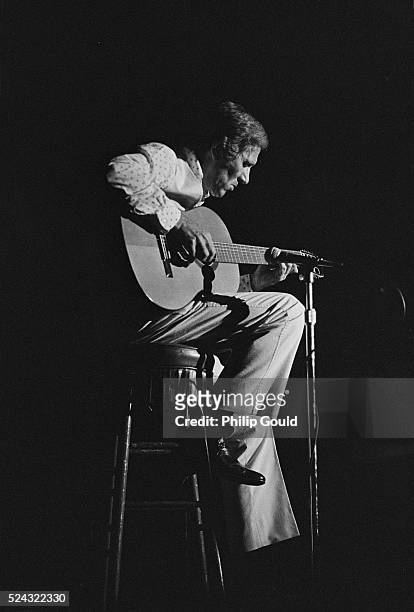 Lit by a stage light, country musician Chet Atkins sits on a stool strumming his acoustic guitar.