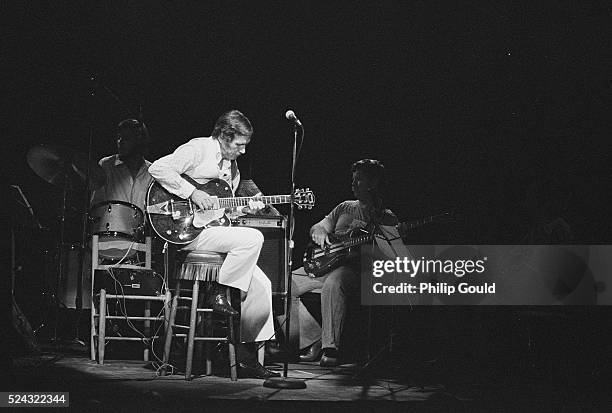 Lit by a stage light, country musician Chet Atkins sits in front of his drummer and bassist, playing his electric guitar.