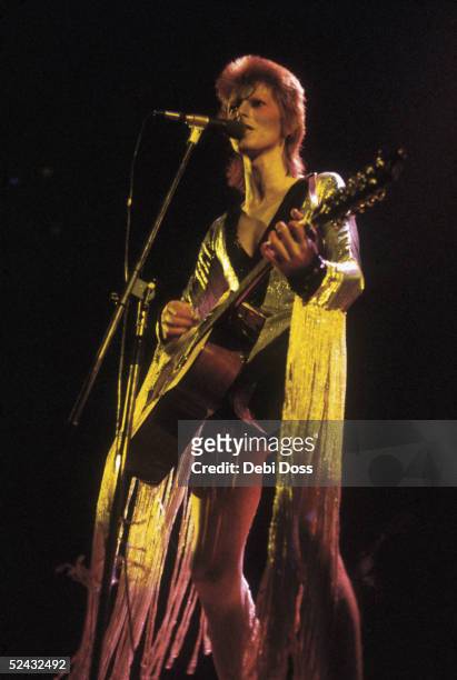 David Bowie performing as Ziggy Stardust at the Hammersmith Odeon, 1973. He is wearing a silver costume with gold tassels by Japanese designer Kansai...