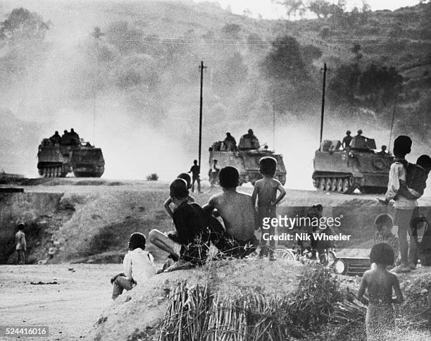 South Vietnamese villagers pause to watch a convoy of US Army armored personnel carriers kick up dust as they drive along a dirt road near Saigon...