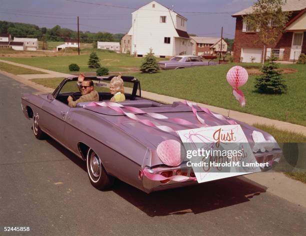 Newly-married couple waves as they drive off with streamers and a 'Just Married' sign on the back of their pink convertible, 1964.