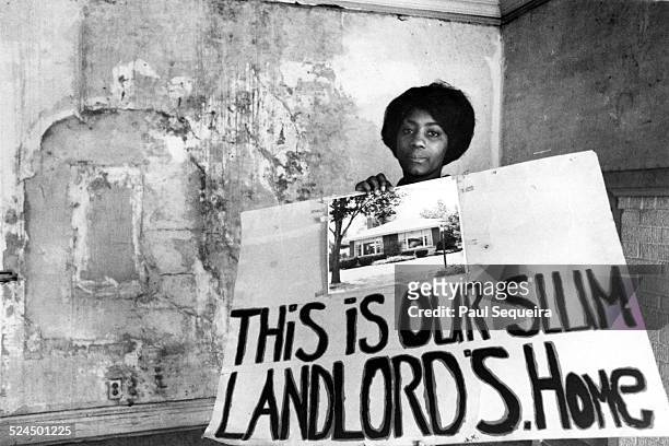 Woman, a resident of the Cabrini Green housing projects, holds up a sign that shows her building landlord's home, Chicago, Illinois, 1960s. The sign...