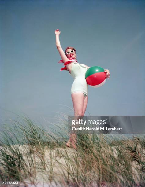 Young woman in a bathing suit holds a beach ball and gestures with one arm on a sand dune, 1956.