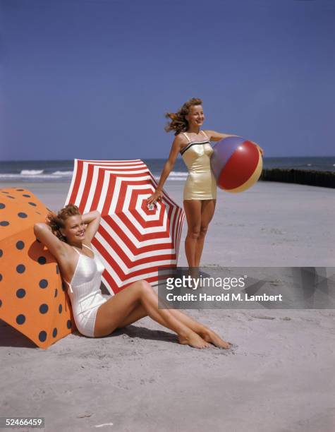 Two women, both redheads, pose on the beach, 1948. One, in a white bathing suit, sits on the sand in front of an orange and black polka-dot umbrella,...