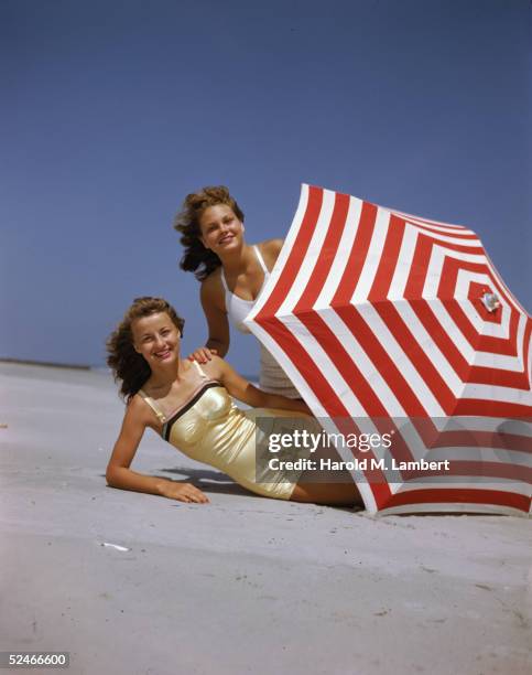 Two women, both redheads, pose behind a red and white umbrella on the beach, 1948. One wears a white bathing suit and the other wears a yellow one.