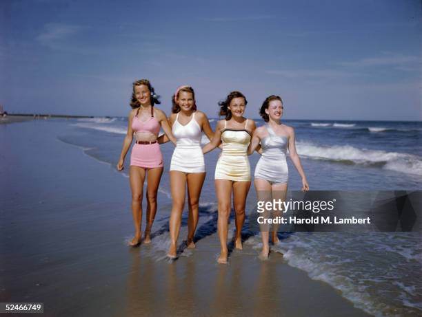 Four young women walk, arm in arm, through the surf, 1949. All four wear bathing suits, from left, a pink two-piece suit, a white halter neck suit, a...