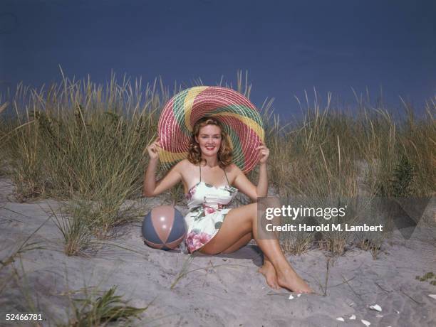 Smiling young woman sits on a sandy ridge, a beach ball by her side, as she holds on to her very large, colorful straw hat, 1956.