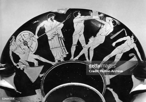 An early Olympiad depicted on an ancient Greek vase or kylix, circa 450 BC. On the left is a competitor in the Hoplitodromos, wearing greaves, a...