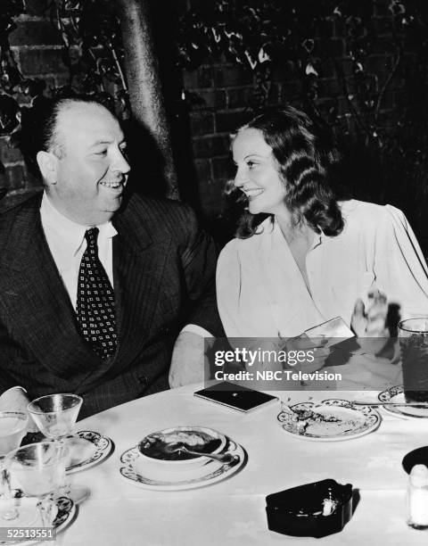 British film director Alfred Hitchcock sits at a restaurant table with American actress Tallulah Bankhead who smiles and taps her cigarette case,...