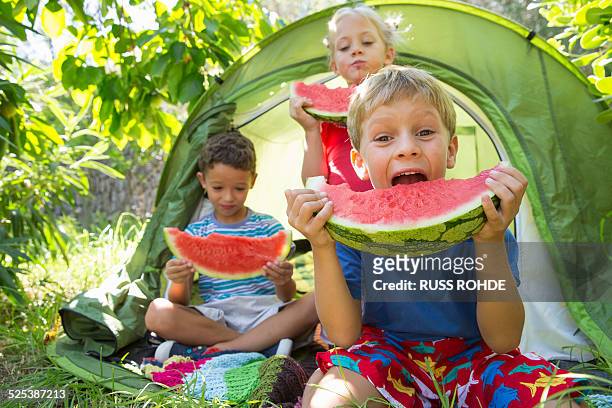 three children eating large watermelon slices in garden tent - children only stock-fotos und bilder