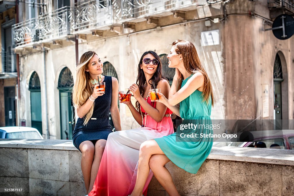 Three young fashionable female friends having cocktails on sidewalk cafe wall, Cagliari, Sardinia, Italy