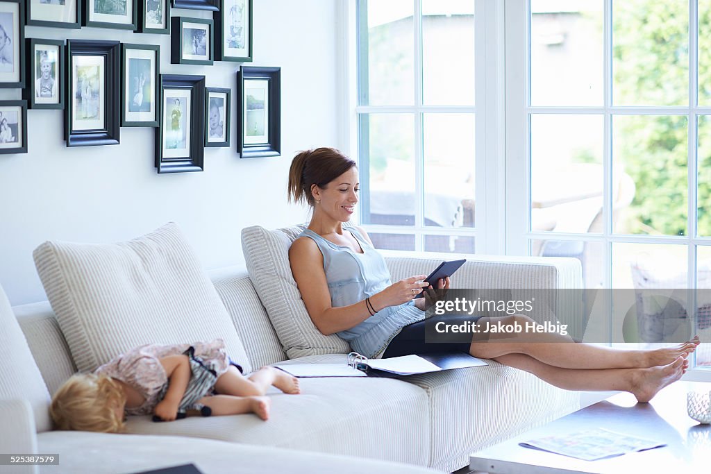 Mid adult woman using digital tablet on living room sofa whilst toddler daughter sleeps