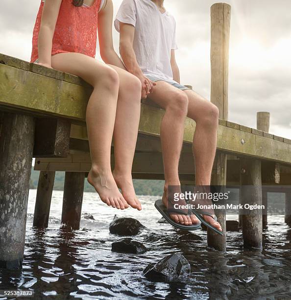 young couple holding hands together and sitting on edge of jetty over lake - windermere stock pictures, royalty-free photos & images