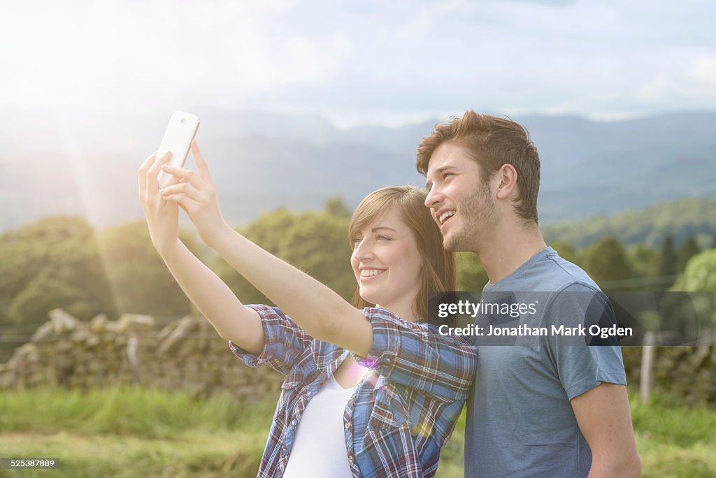 Young couple taking self portrait on mobile phone in countryside under sunny sky