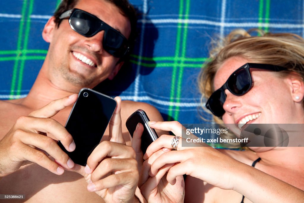 Young couple lying on beach towel using smartphones