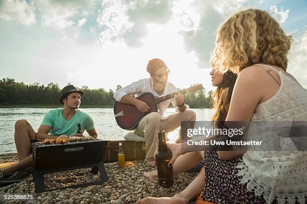 young man sitting by lake with friends playing guitar - strandparty stock-fotos und bilder