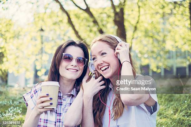 two young female friends drinking coffee and listening to music on headphones in park - sean malyon stock pictures, royalty-free photos & images