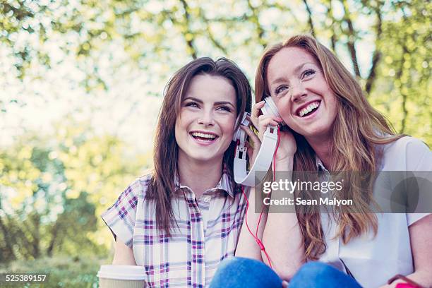 two young female friends drinking coffee and sharing music on headphones in park - sean malyon stock-fotos und bilder