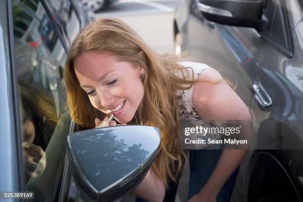 young woman applying lipstick whilst looking in car wind mirror - sean malyon stockfoto's en -beelden