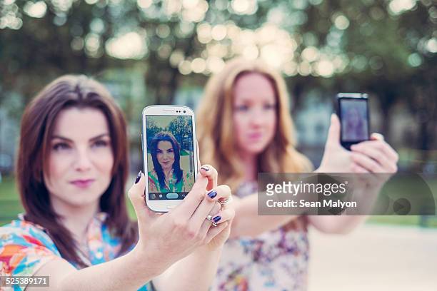 two young female friends taking selfie on smartphones in unison - sean malyon stockfoto's en -beelden