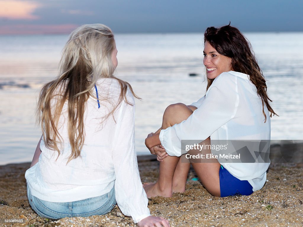 Two young women friends sitting chatting on beach at sunset, Williamstown, Melbourne, Australia