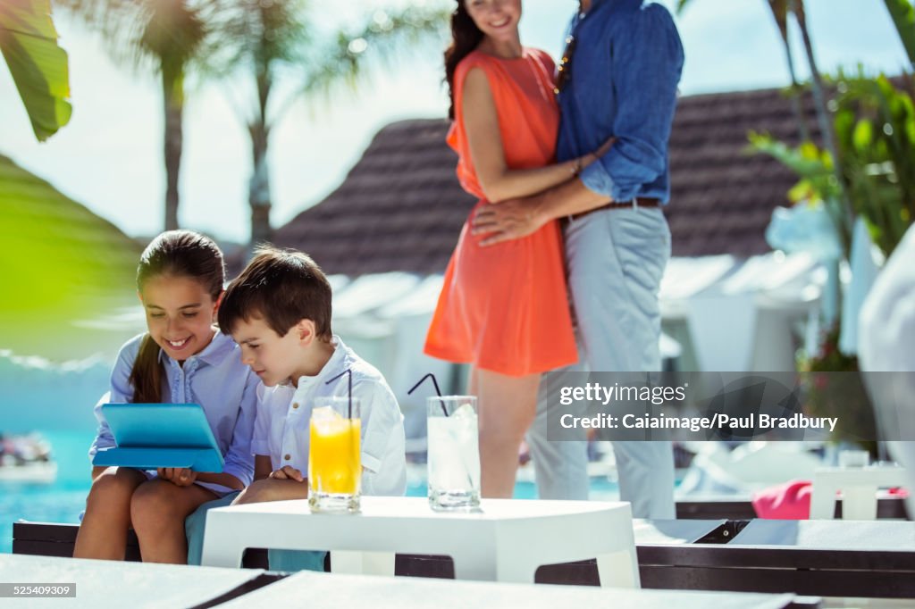 Fratello e sorella che usano il tablet digitale dalla piscina del resort, i genitori che abbracciano in background