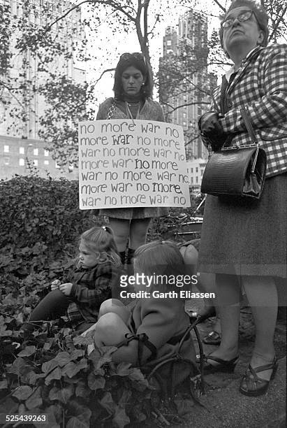 An antiwar demonstrator holds a sign as she looks at a pair of young girls seated in an ivy patch in Bryant Park during a Vietnam War protest, New...