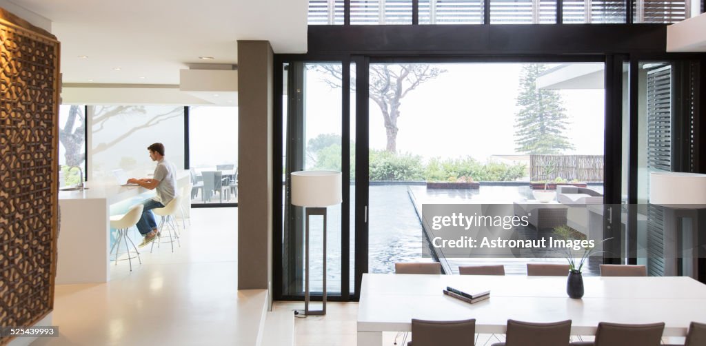 Young man using laptop in modern kitchen, dining area with patio door and swimming pool in foreground