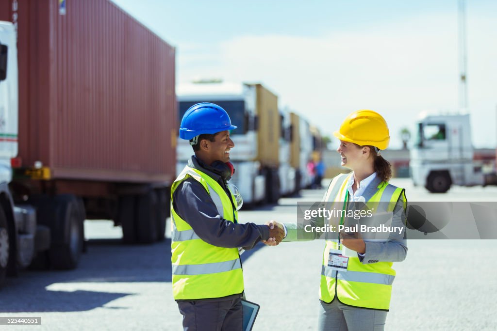 Worker and businesswoman shaking hands near trucks and cargo containers