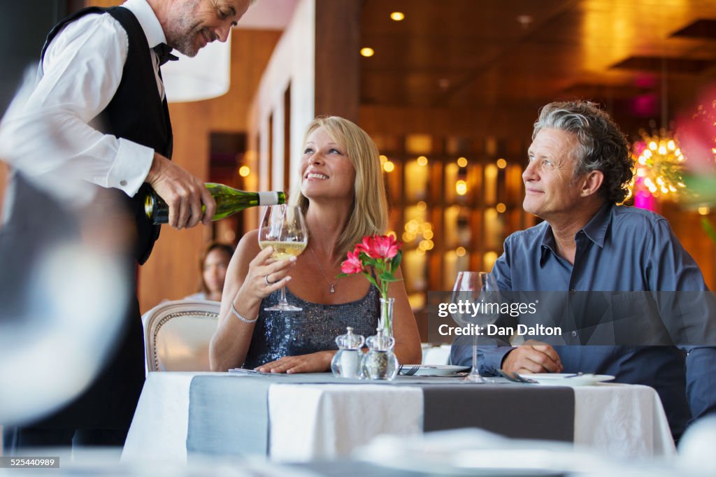 Mature couple sitting at restaurant table, waiter pouring white wine into womans glass