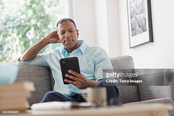 usa, new jersey, jersey city, man sitting on sofa at home and using digital tablet - mature man using digital tablet stock pictures, royalty-free photos & images