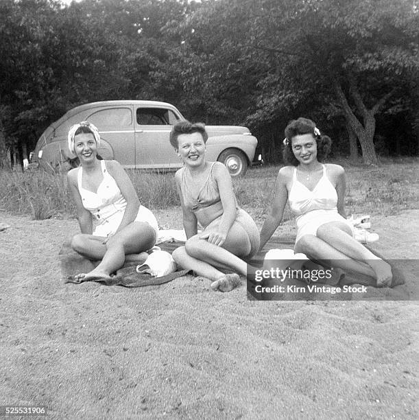 Three young women lounge on beach towels on the sand with their car in the background.